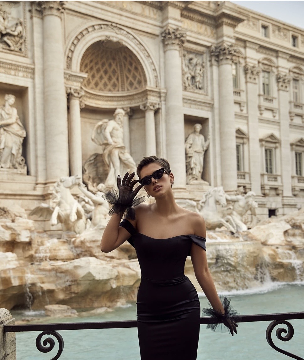 Woman in black dress and sunglasses standing in front of ornate fountain with sculptures, waving hand, holding railing, outdoors.