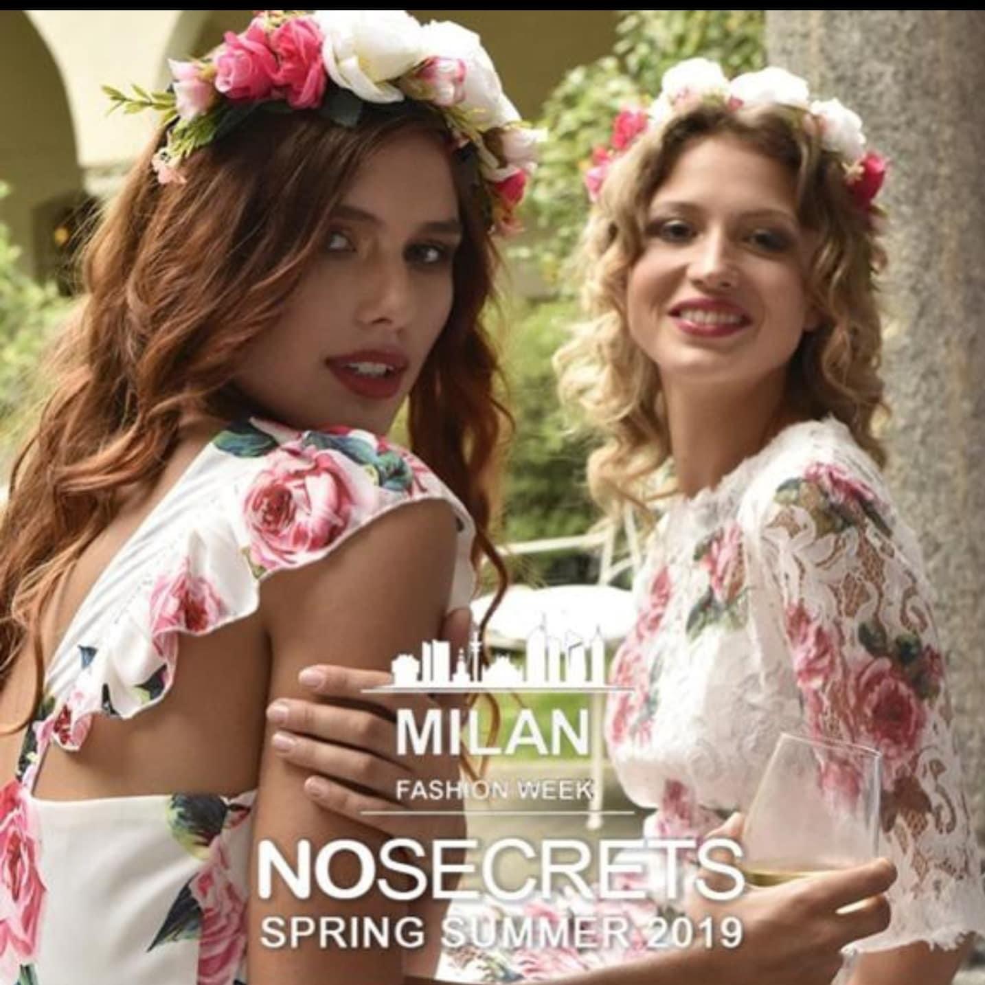 Two women wearing floral dresses and flower crowns, smiling outdoors during spring or summer, holding drinks.