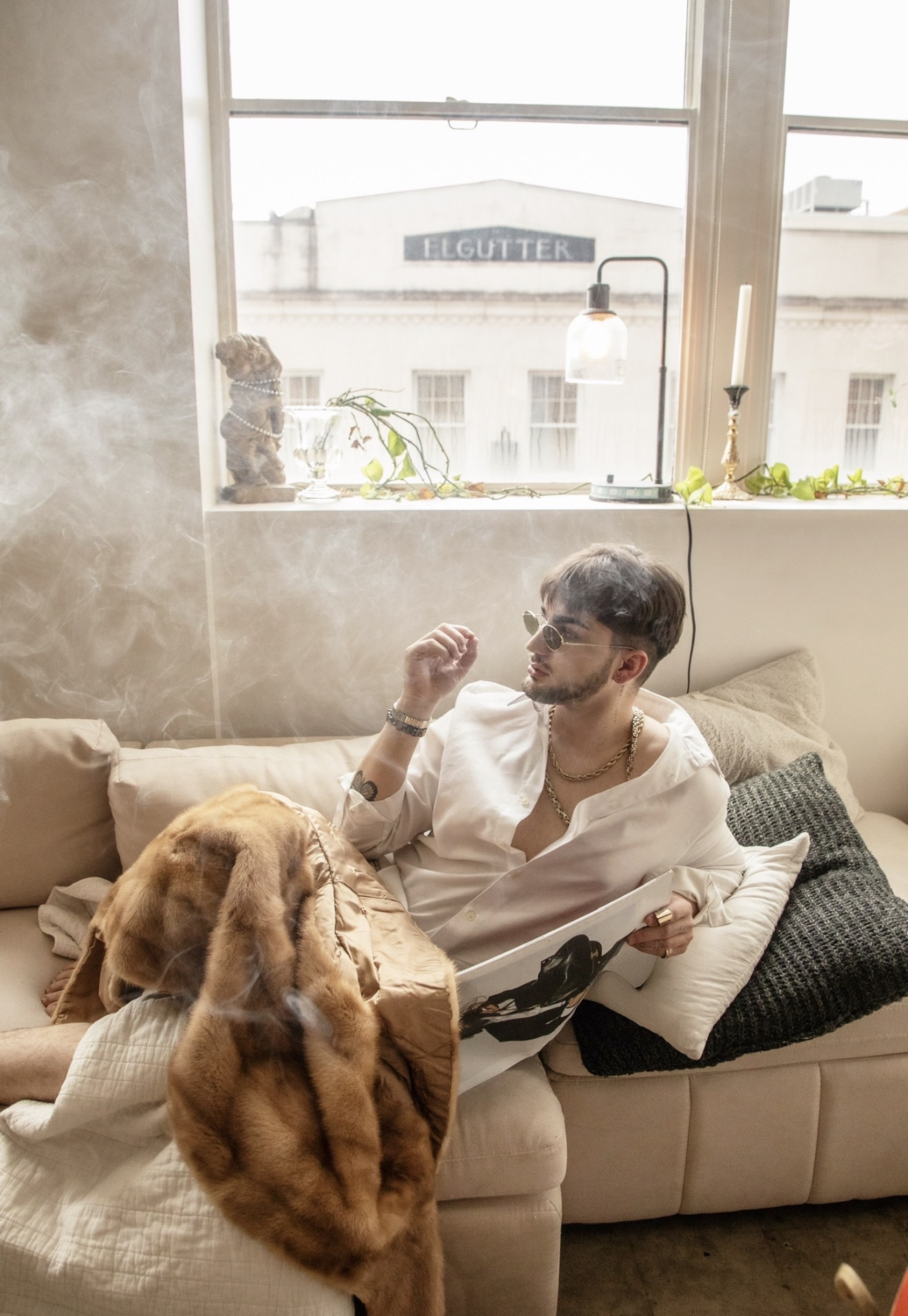 Man with dark hair and beard relaxing on a sofa with a dog, in a room with large window and plants.