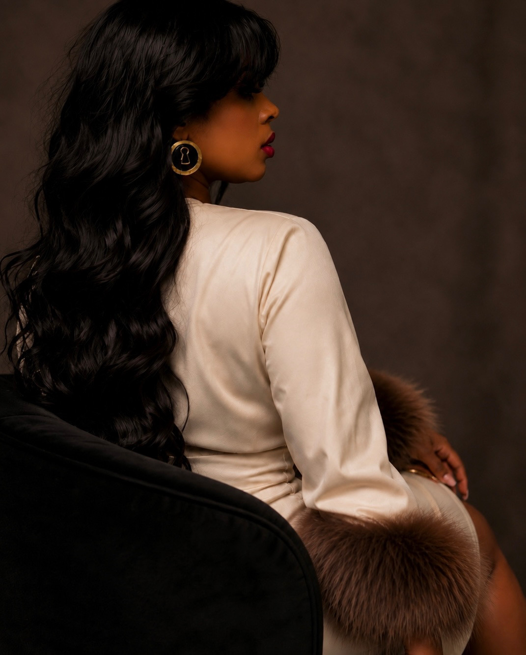 Side view of a woman with long black hair, wearing a white top and large earrings, sitting with a fur stole.