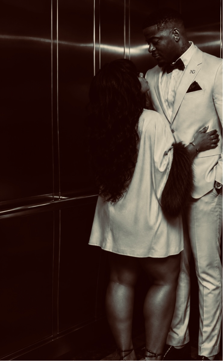 A man in a white suit and bow tie stands close to a woman with long hair in an elevator, holding her arm.