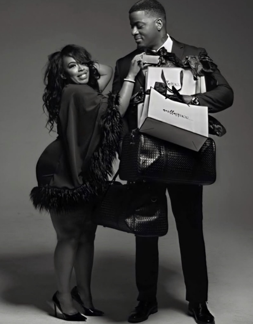 A man and woman dressed elegantly, holding shopping bags, smiling, standing close together, woman leaning on man, in black and white.