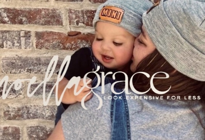 Child wearing a gray beanie with orange text being held by a person in a gray hoodie, against a brick wall.