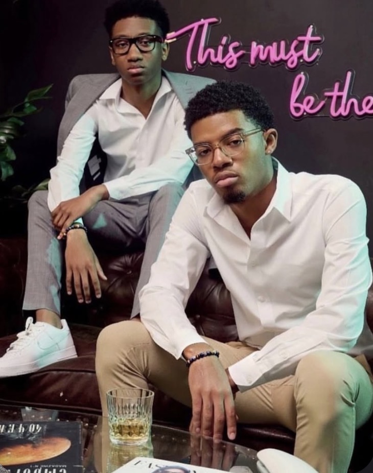 Two men sit on a black leather couch, one in a white shirt and beige pants, the other in a white shirt and gray pants, with a neon pink sign in the background reading 'This must be the'.