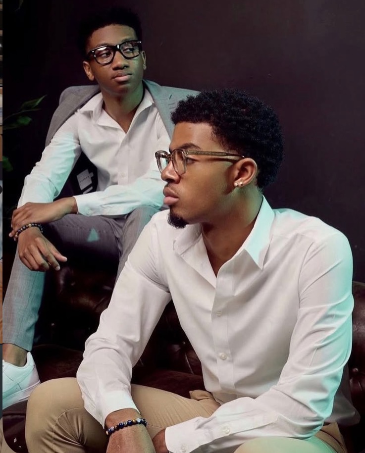 Two young men with glasses sit on a dark background, wearing white shirts, one looking to the side, the other looking forward.