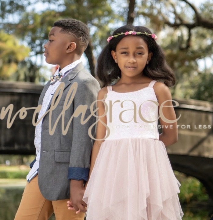 A young girl and boy holding hands outdoors, girl in a pink dress and girl in a gray blazer, trees in background.