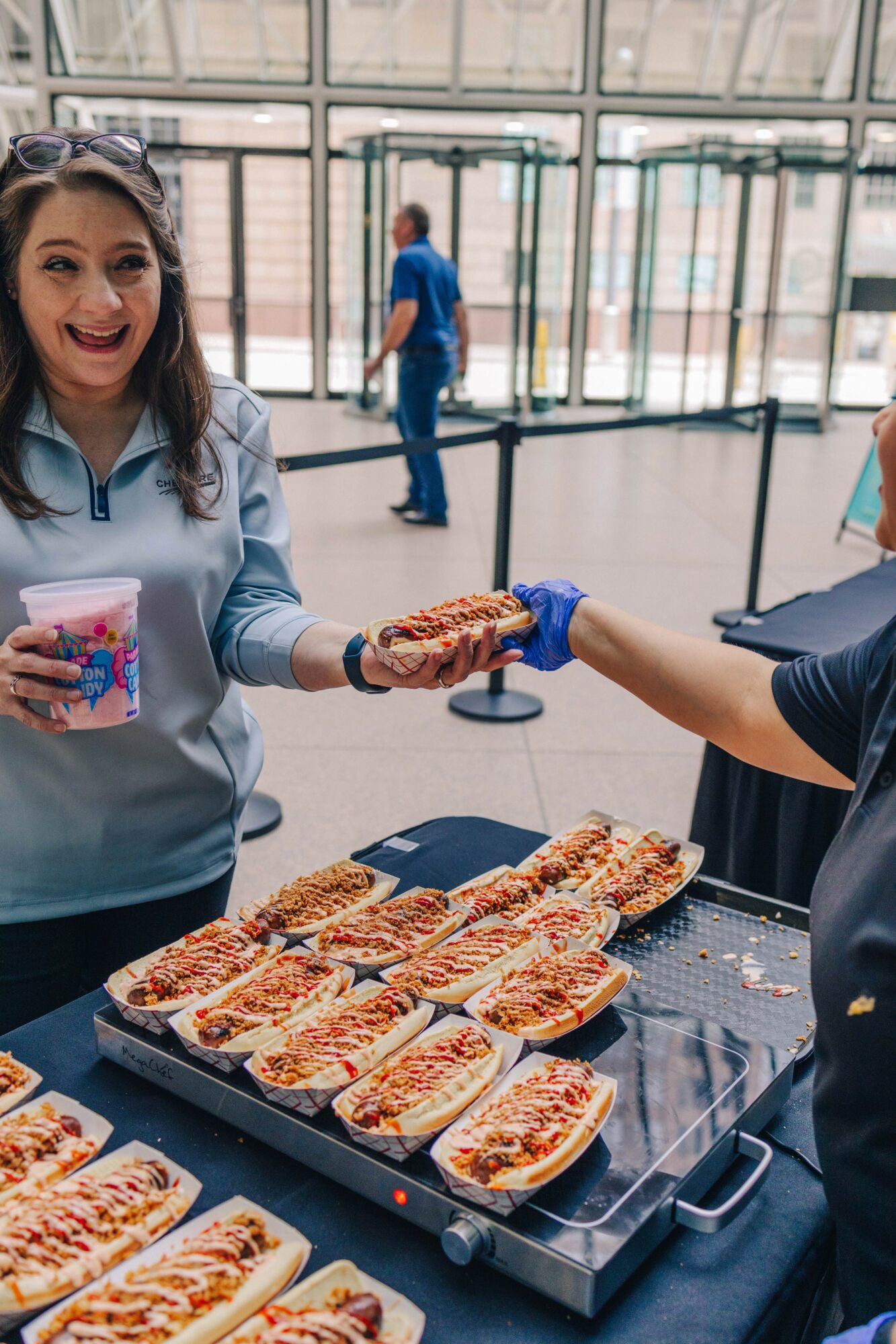 Woman smiling and holding a drink, receiving a hot dog from a server at a food stand, with trays of hot dogs on the table.