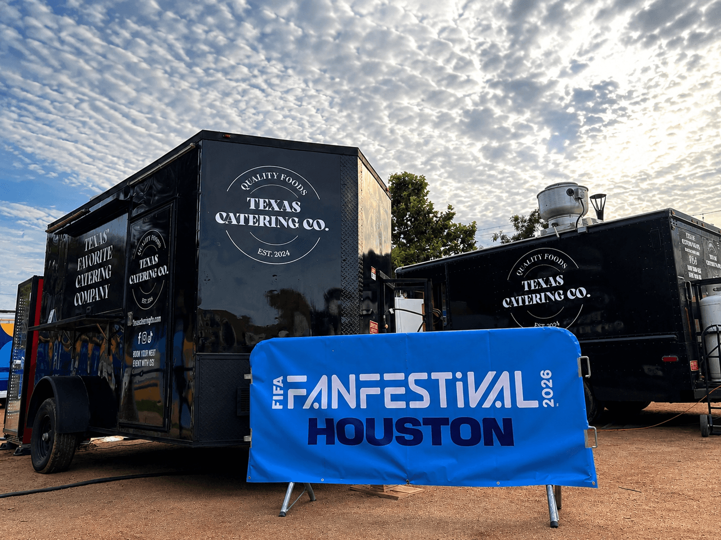 Two black food trucks with Texas Catering Co. logos parked outdoors under a partly cloudy sky, with a blue Houston Fan Festival banner in front.