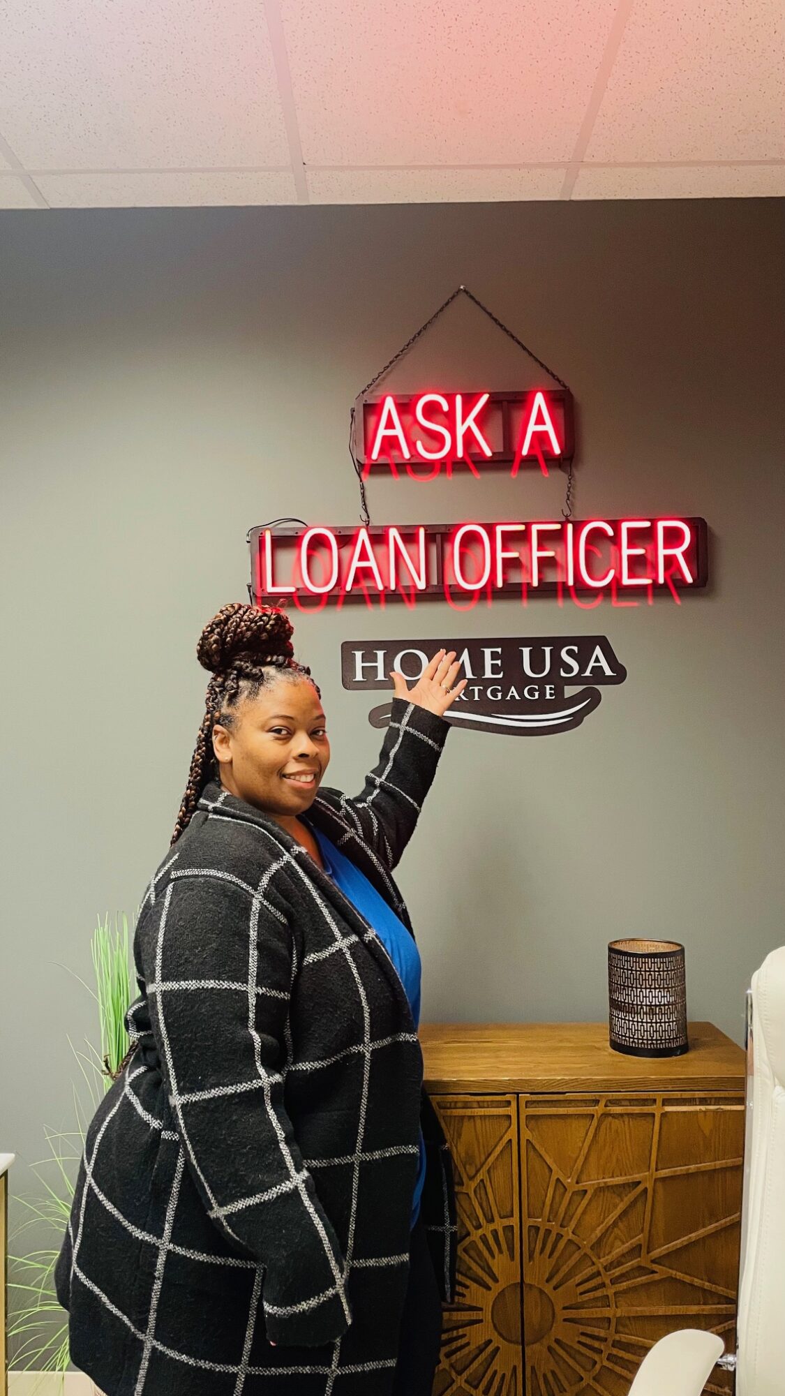 Woman pointing at a neon sign that reads 'Ask a Loan Officer' in a room with a wooden cabinet and a decorative item.