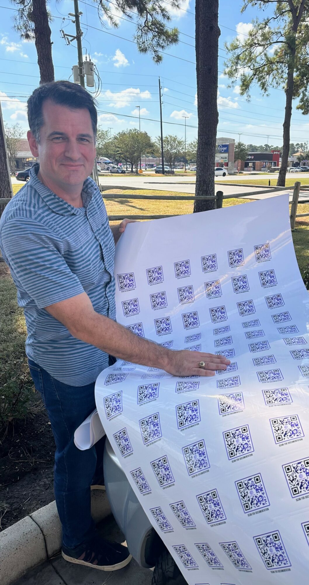 Man in striped shirt holding a large sheet with printed patterns outdoors, trees and a parking lot in background.