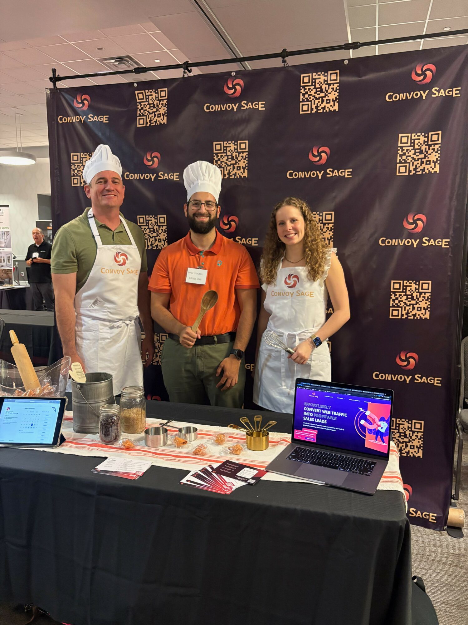 Three people stand behind a table with a laptop and promotional materials, in front of a backdrop with logos and QR codes.