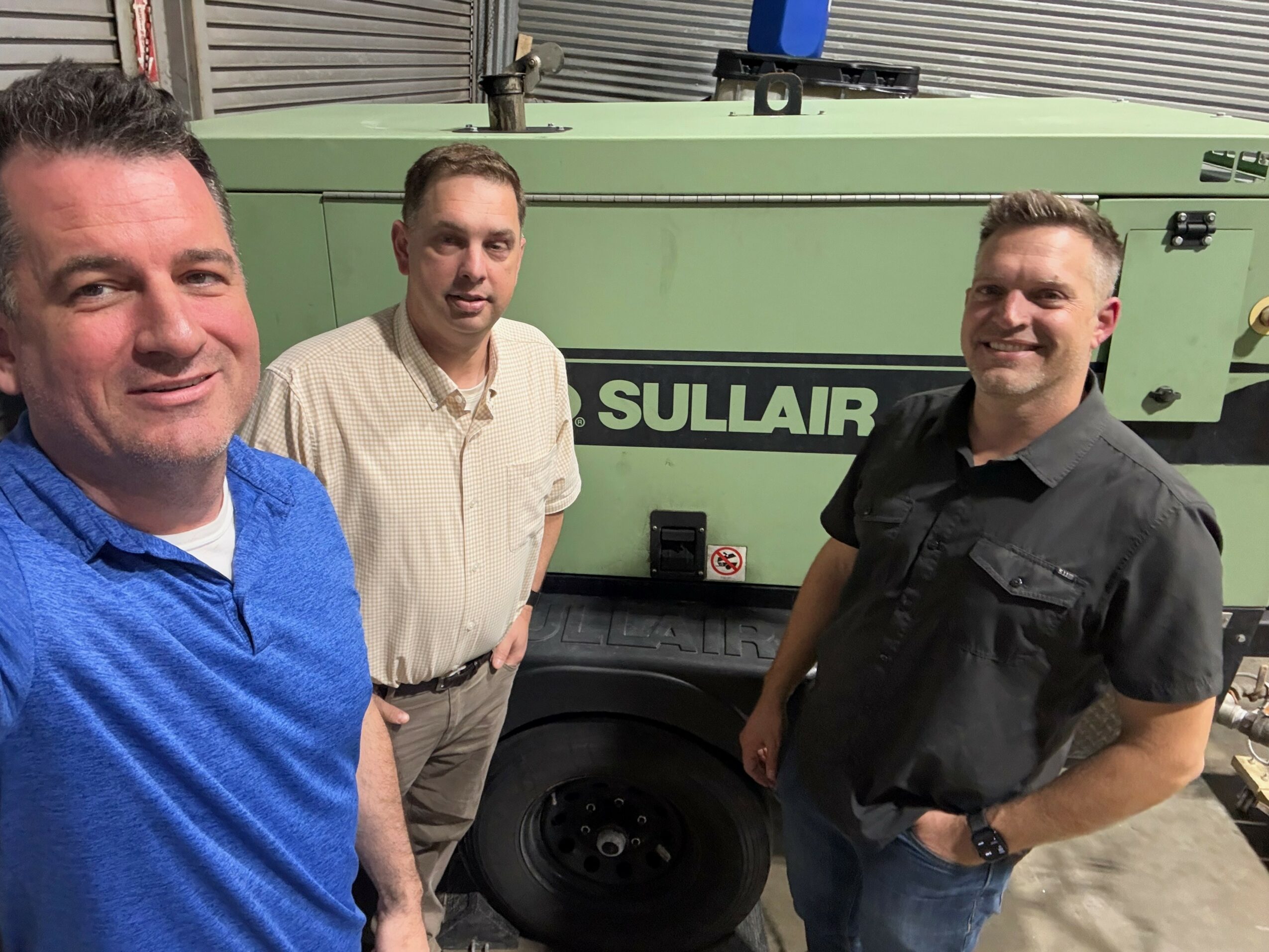 Three men standing in front of a large green Sullair industrial machine, smiling, in an indoor setting.