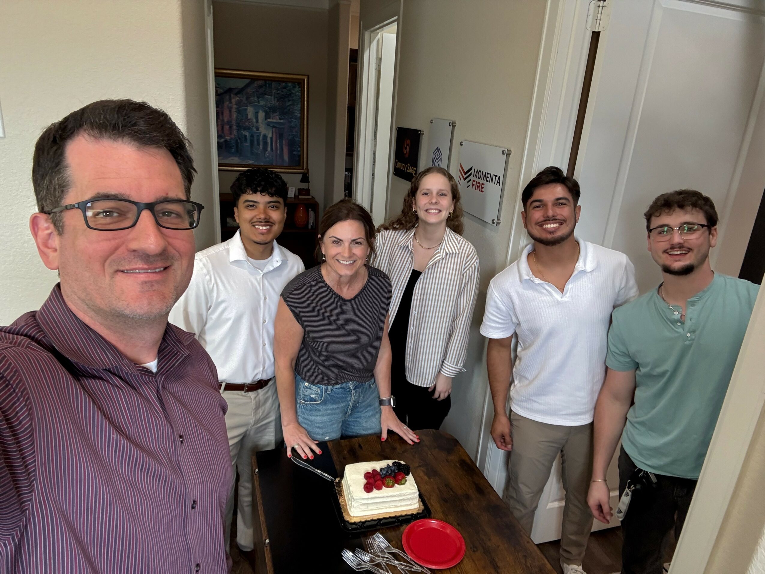 Group of six people standing around a table with a cake, smiling indoors.