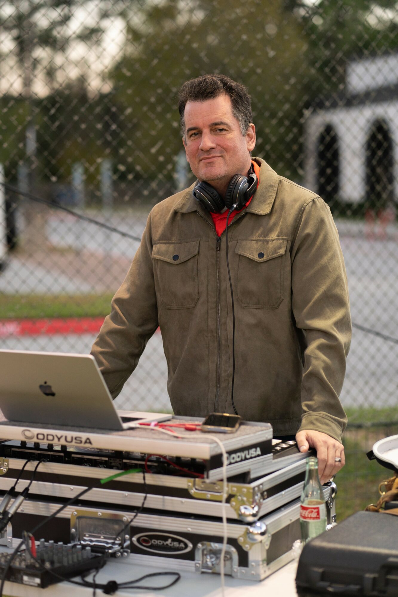 Man wearing headphones and a jacket stands behind DJ equipment outdoors, with a chain-link fence in the background.