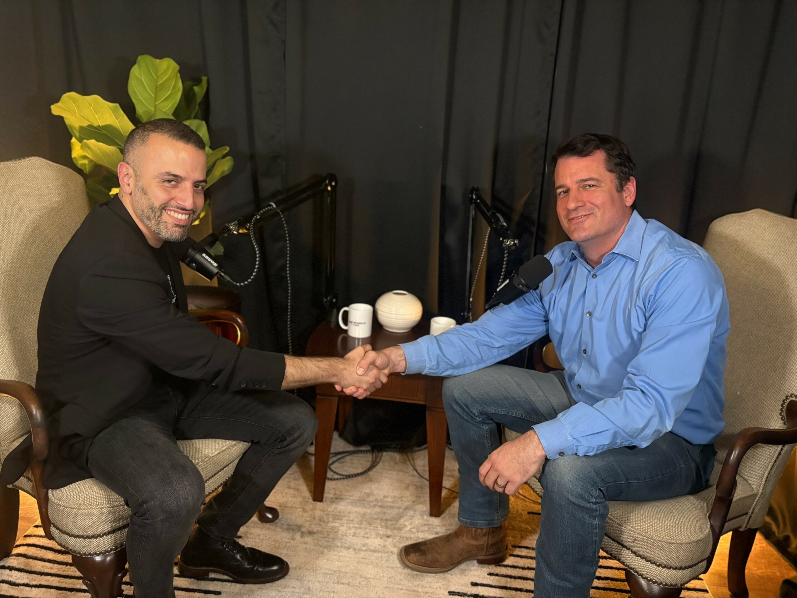 Two men shake hands while sitting in chairs, smiling, with microphones and a small table between them, in a room with dark walls and plants.