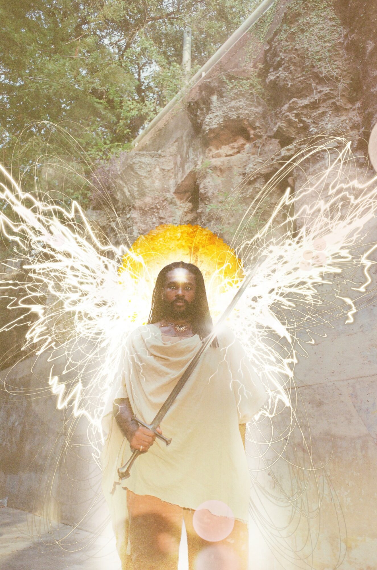 Person dressed as Jesus with wings and a halo, holding a staff, outdoors near rocks and trees.