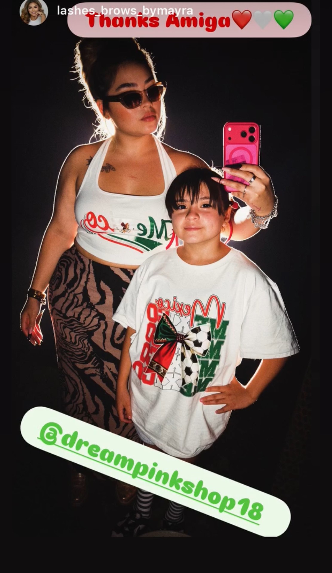 Two people taking a mirror selfie, one adult woman and one child, both wearing white shirts with colorful designs, dark background.
