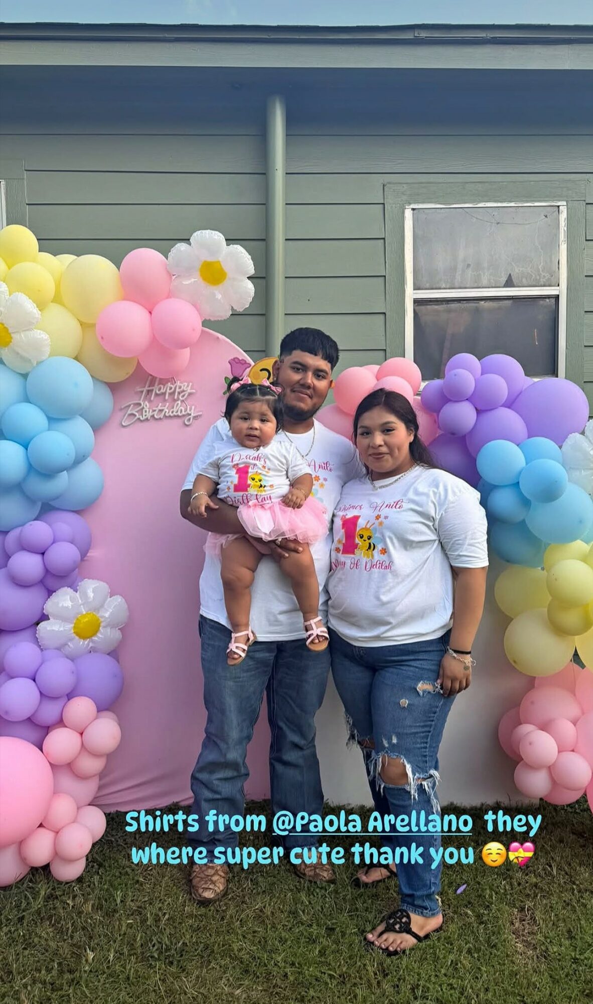 Family of three standing outside in front of colorful balloon decorations, holding a birthday cake, smiling.