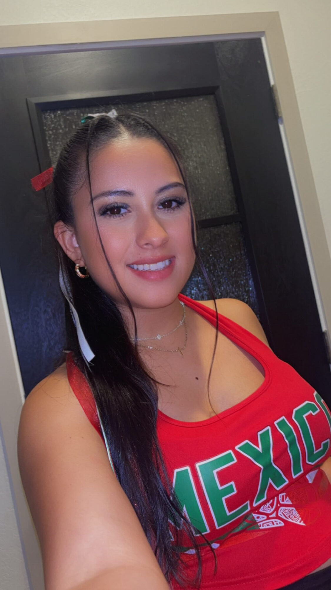 Young woman with long dark hair, wearing a red top with green text, smiling, indoors.