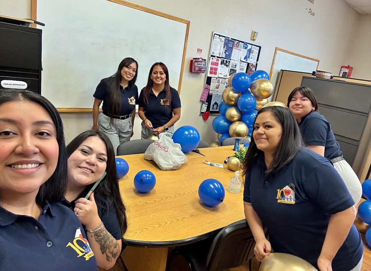 Six women in navy shirts gathered around a table decorated with blue and gold balloons in a classroom.