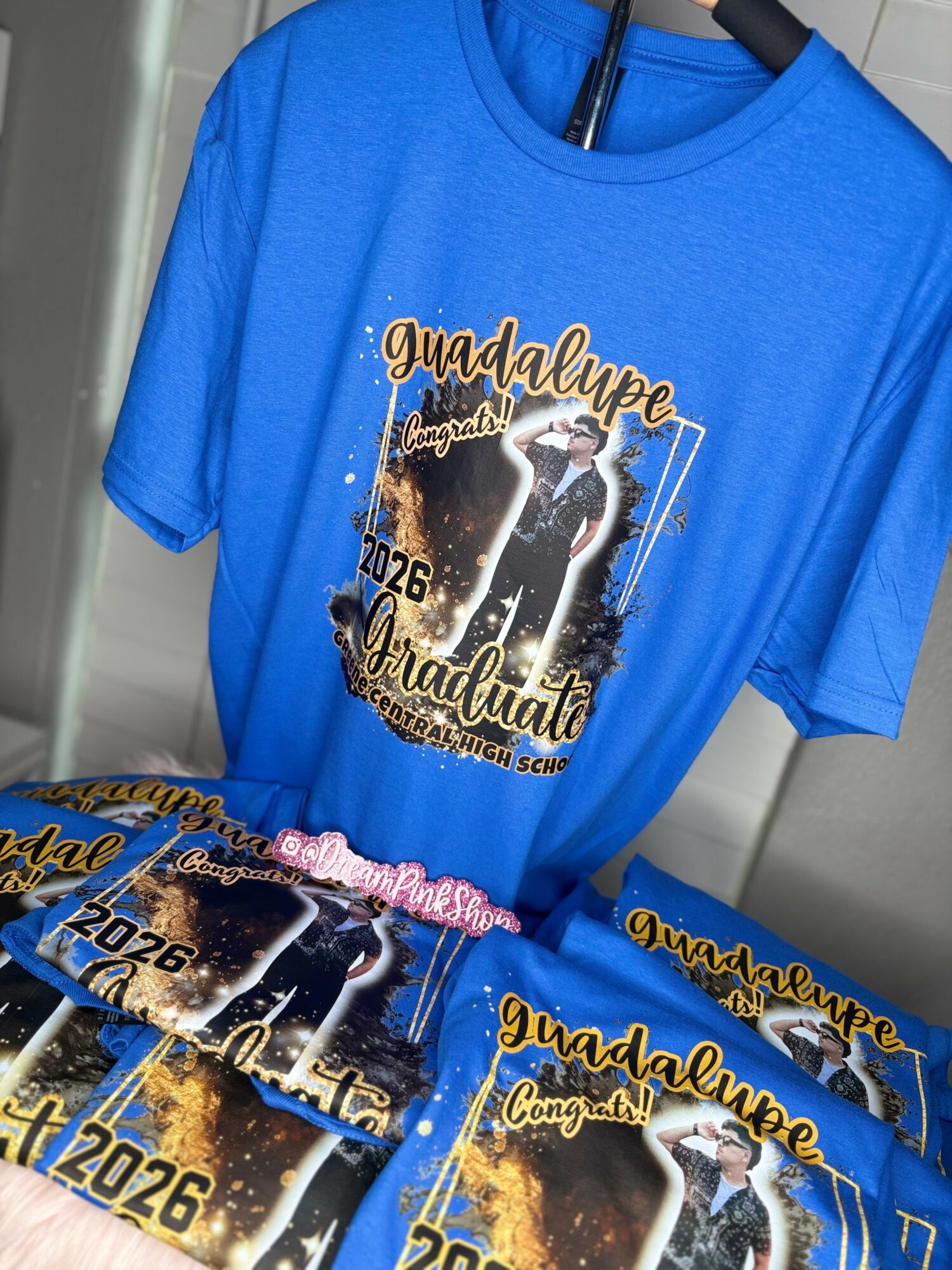 Blue graduation t-shirt with a graphic of a person in a cap and gown, text, and decorative elements, hanging on a hanger.