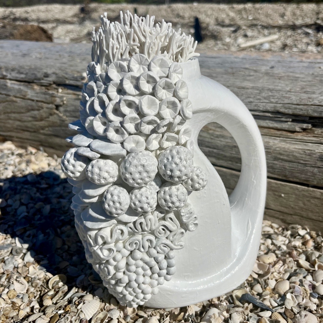 White ceramic mug with textured, sculpted fruit and plant designs on the front, placed outdoors on pebbles with a wooden log background.
