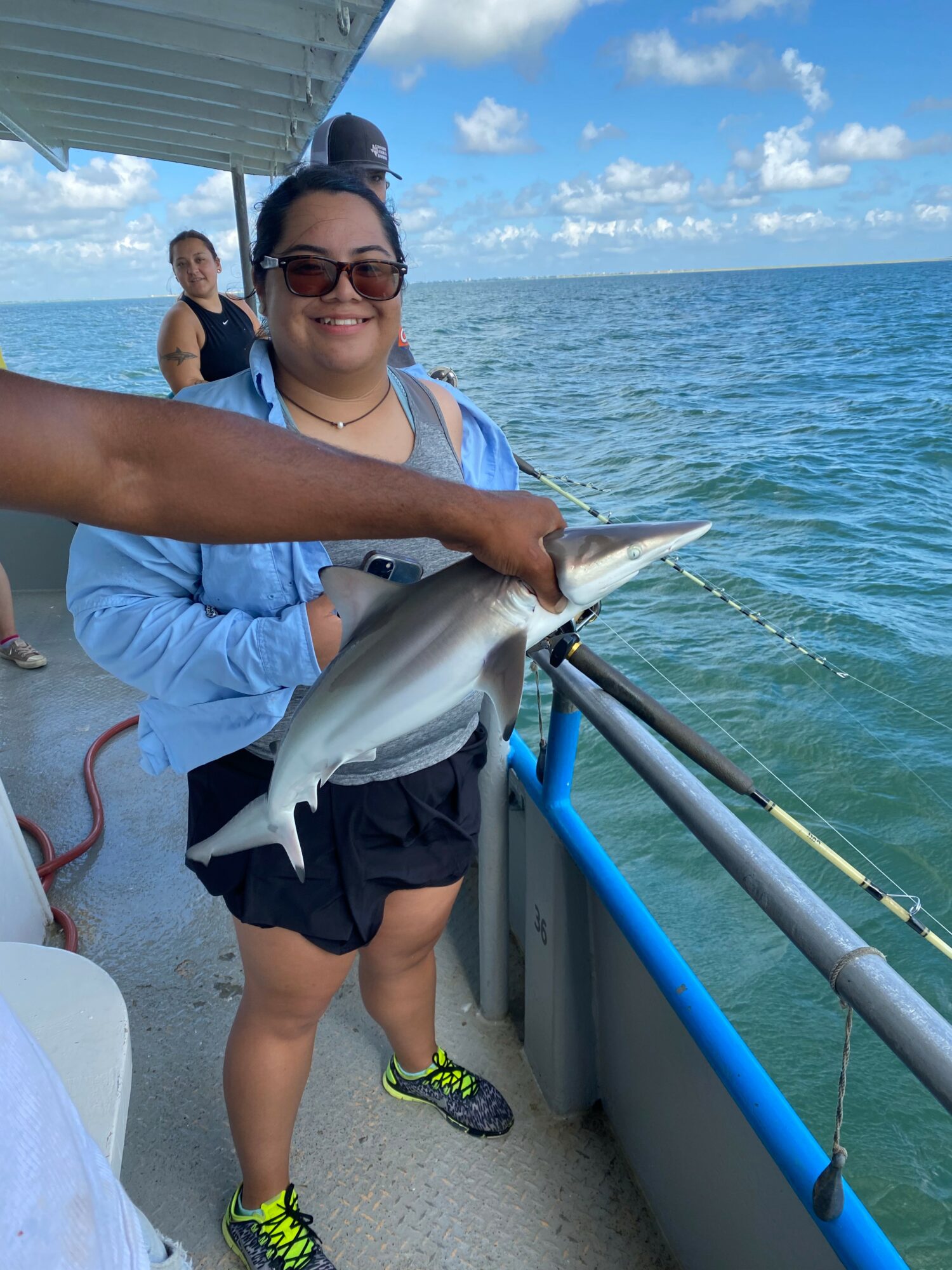 Woman holding a fish on a boat with ocean and sky in background, smiling, wearing sunglasses and casual clothing.