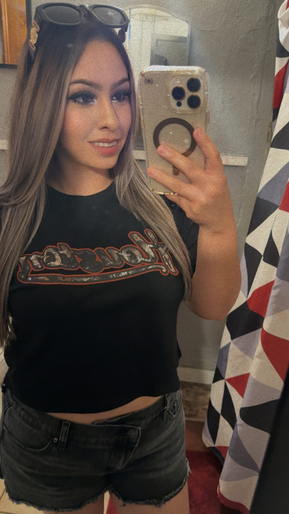 Young woman taking a mirror selfie with sunglasses on her head, wearing a black shirt and shorts, holding a phone with a patterned case.