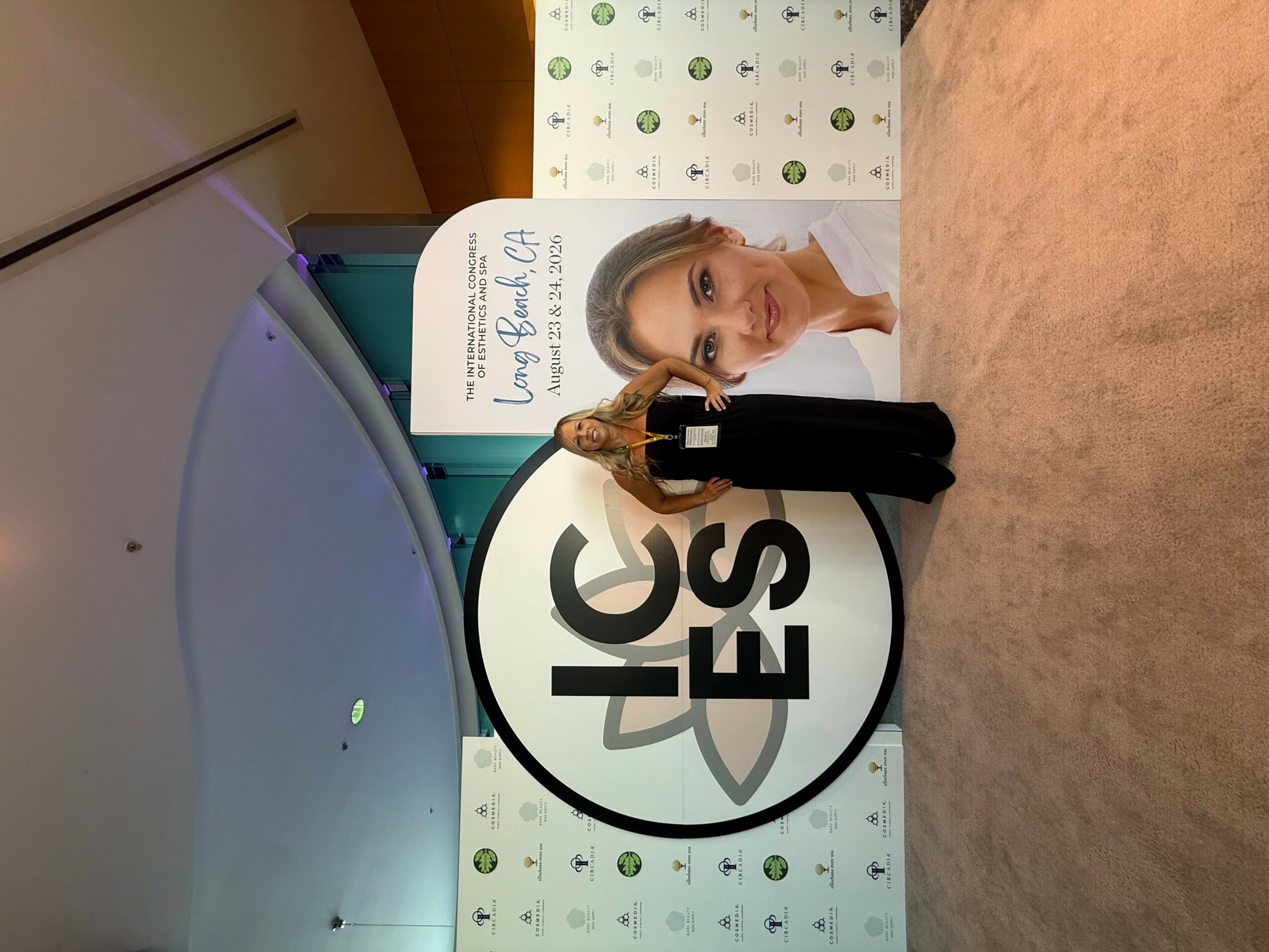 Person with long hair and white shirt at a booth with a large logo and a backdrop with small icons, facing camera.