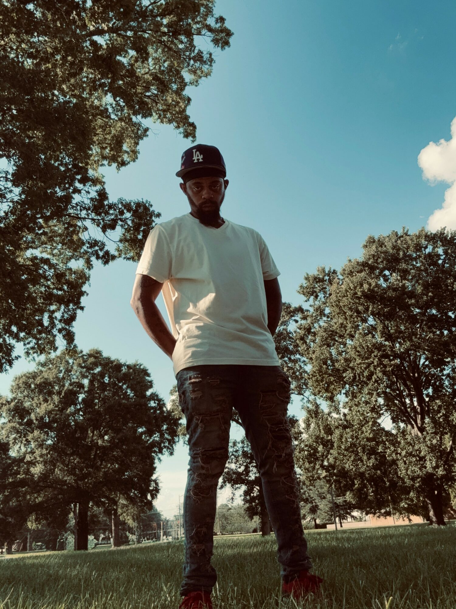 Person standing outdoors in a park with trees and blue sky, wearing a white t-shirt, dark pants, and a cap.