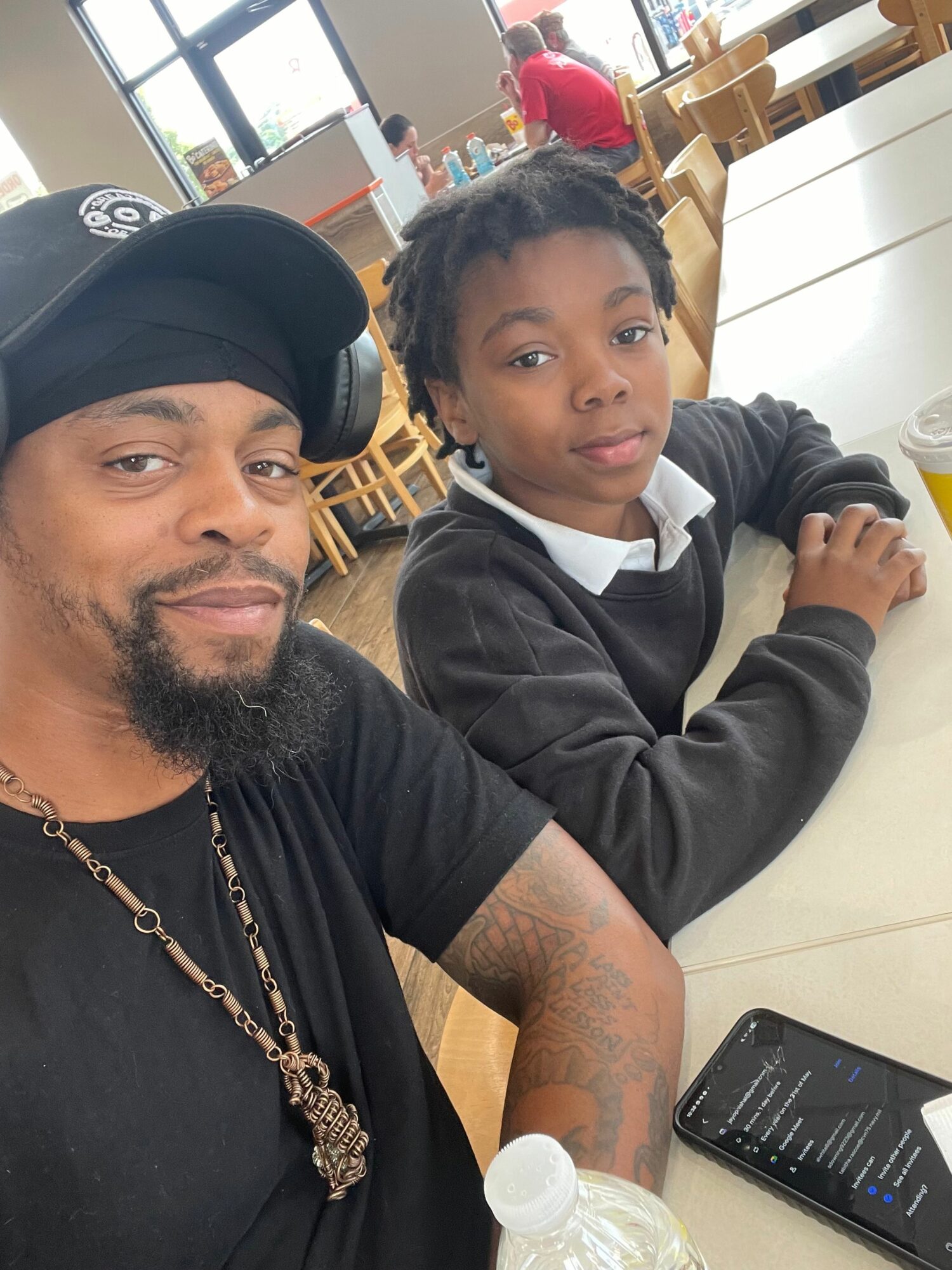 Two people sitting at a table, one with a black hoodie and the other with a black hat, smiling, with a smartphone nearby.