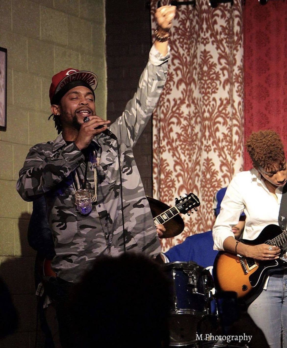 Performer singing into microphone with raised fist, holding guitar, on stage with patterned curtains, wearing camouflage jacket and cap.