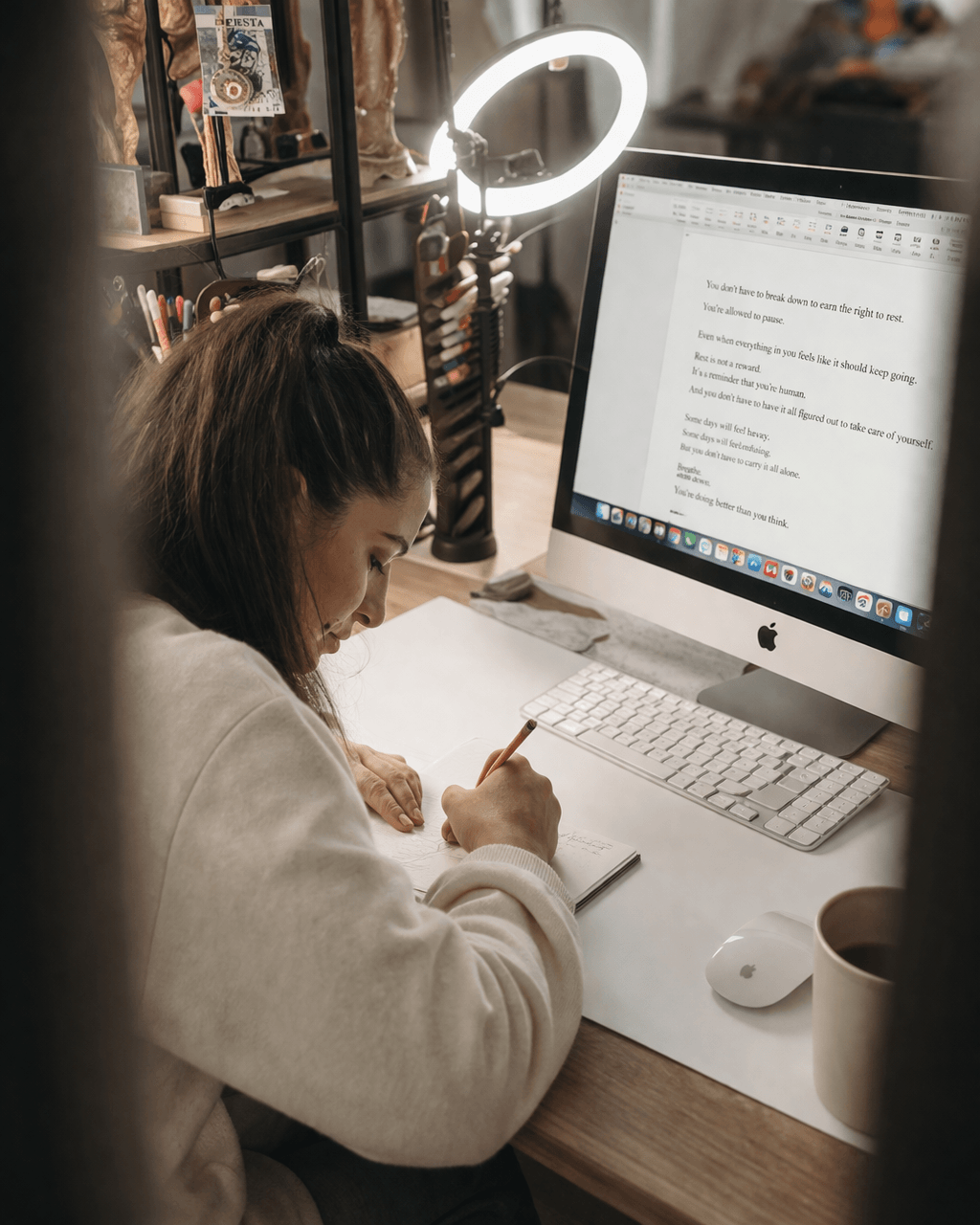 Person writing notes at desk with computer, ring light, and books in background.