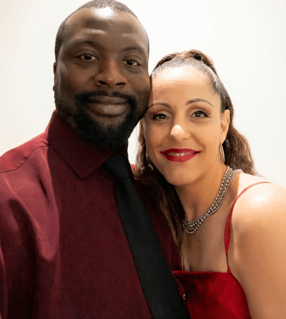 Two people smiling, close-up portrait, woman with red dress and necklace, man with dark shirt and tie, plain background.