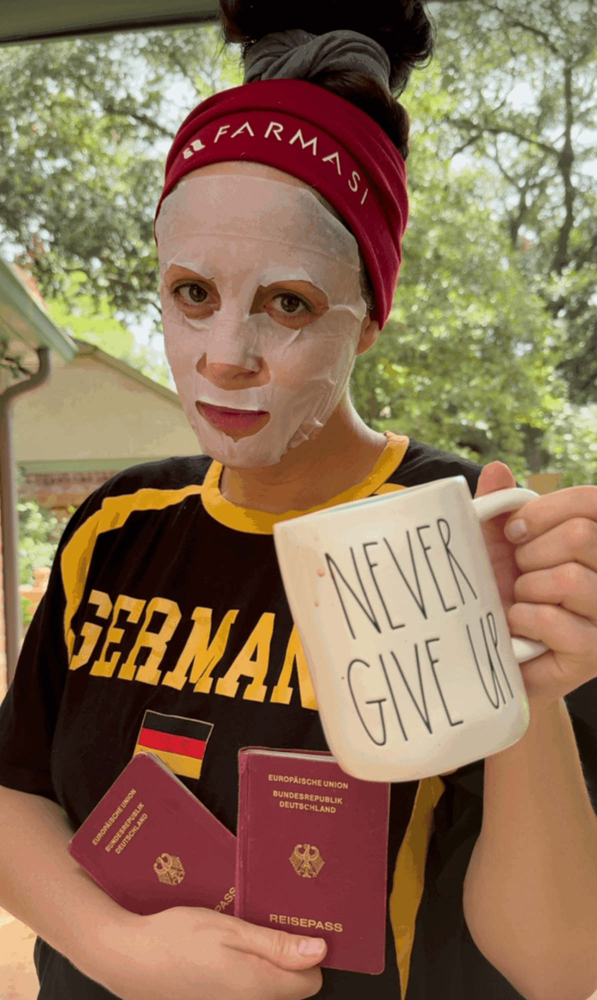 Person with a face mask, holding a mug that says 'Never Give Up,' and two passports, outdoors with trees in background.