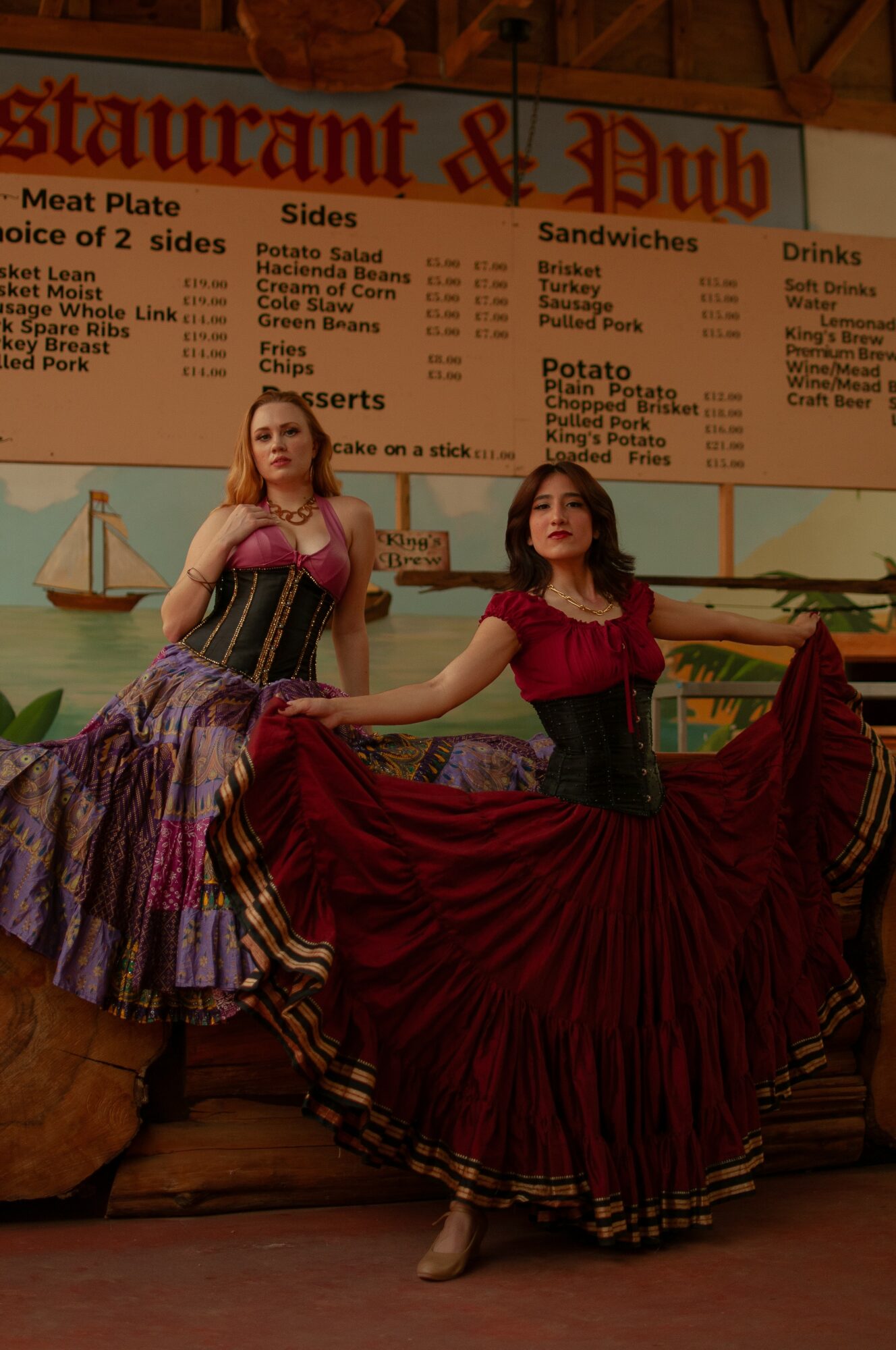 Two women in colorful dresses dance indoors, holding hands, with a restaurant menu in the background.