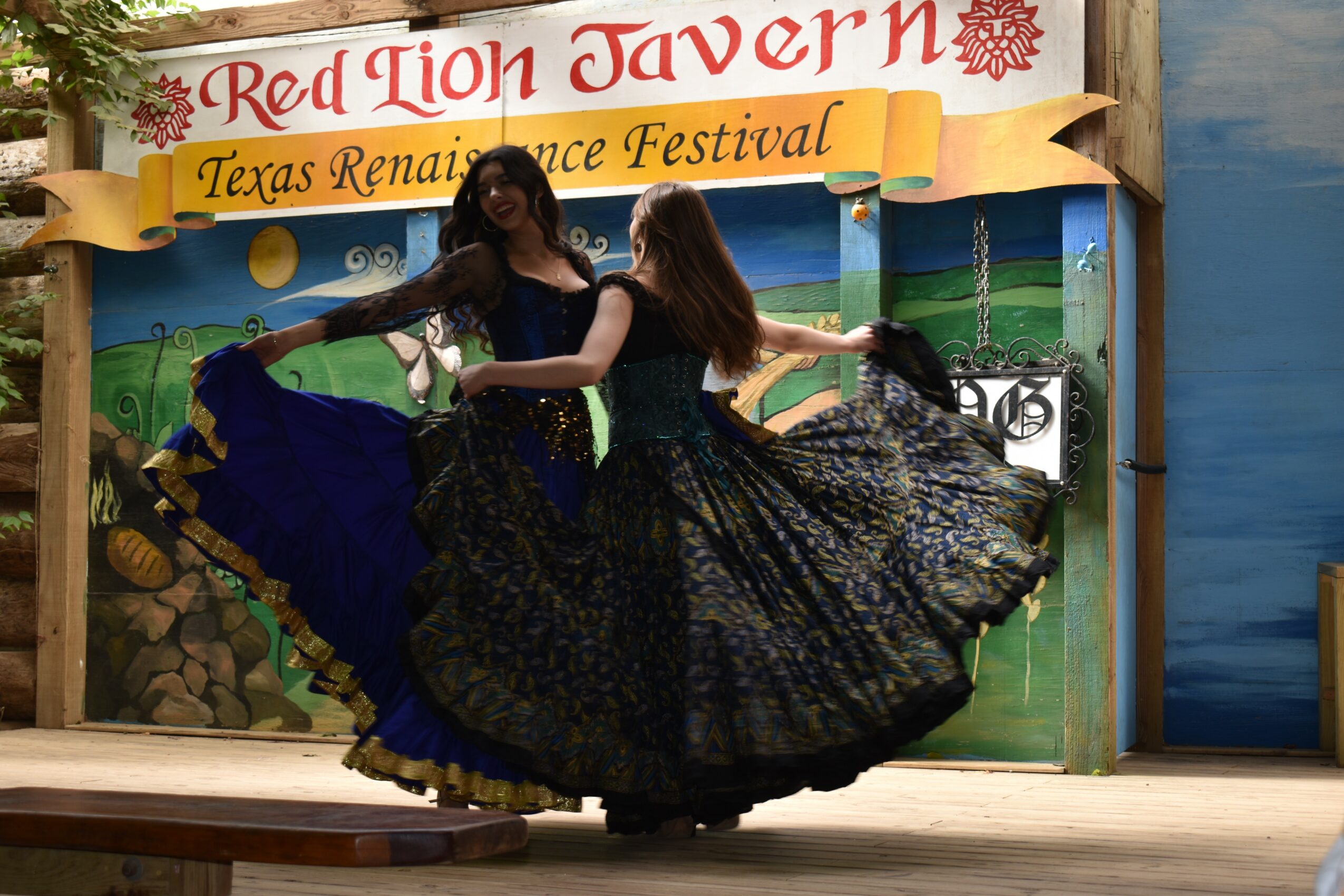 Two women in traditional dresses dance on stage at Texas Renaissance Festival, with a colorful backdrop and a sign reading 'Red Lion Tavern'.