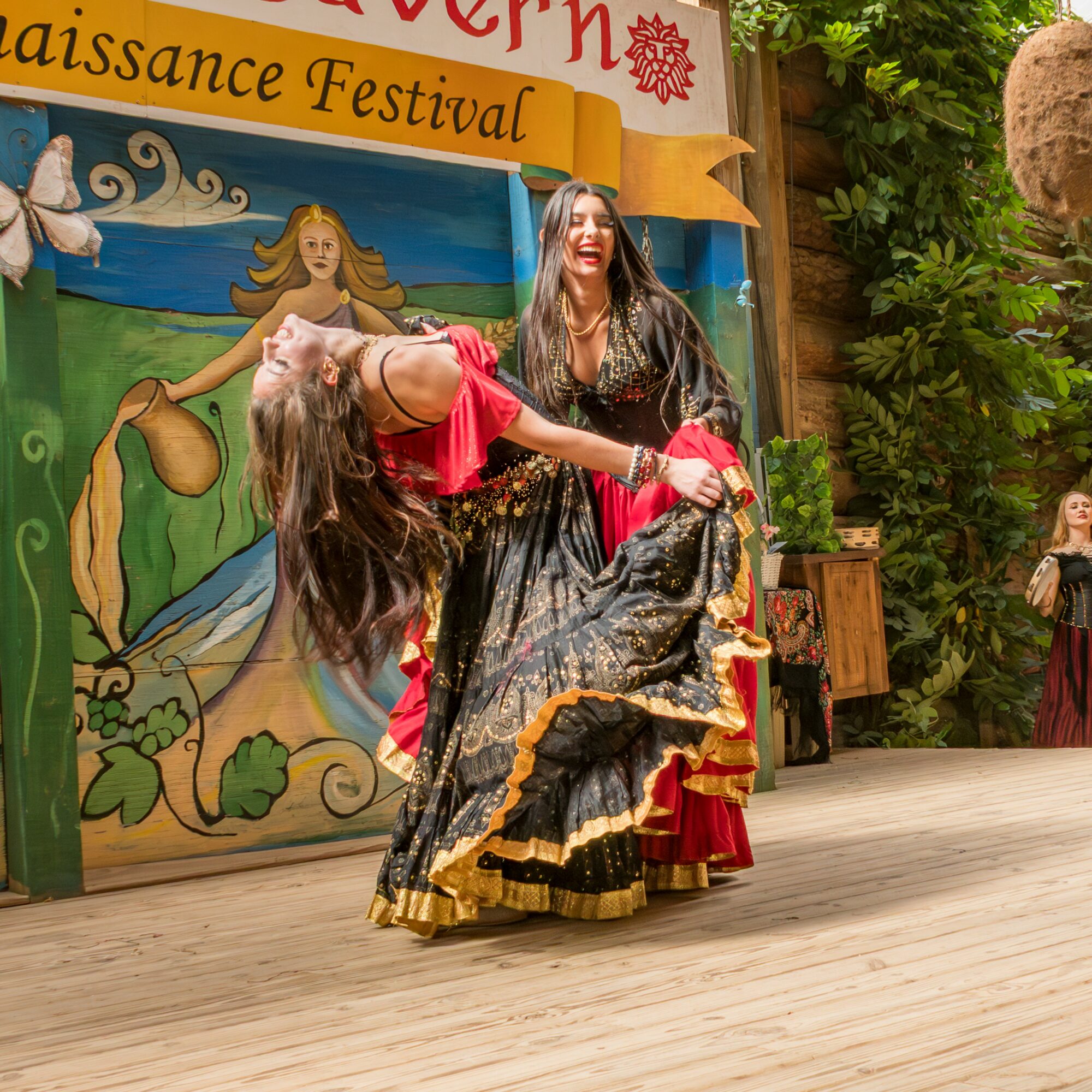Two women dancing on stage, one in a black and gold dress, the other in a red dress, with a colorful backdrop. Word banner at top.