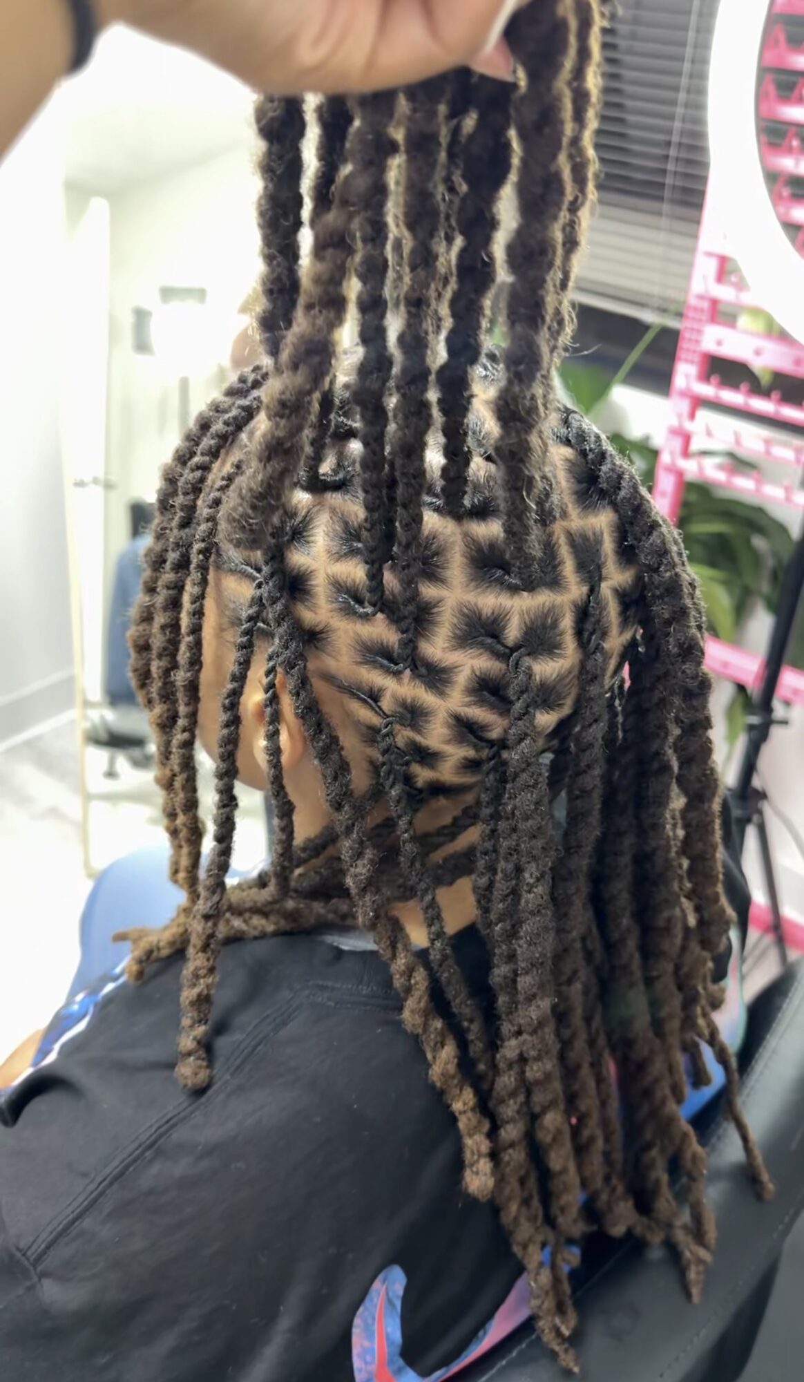 Person with long, styled dreadlocks sitting in a salon chair, facing away, with salon equipment in the background.