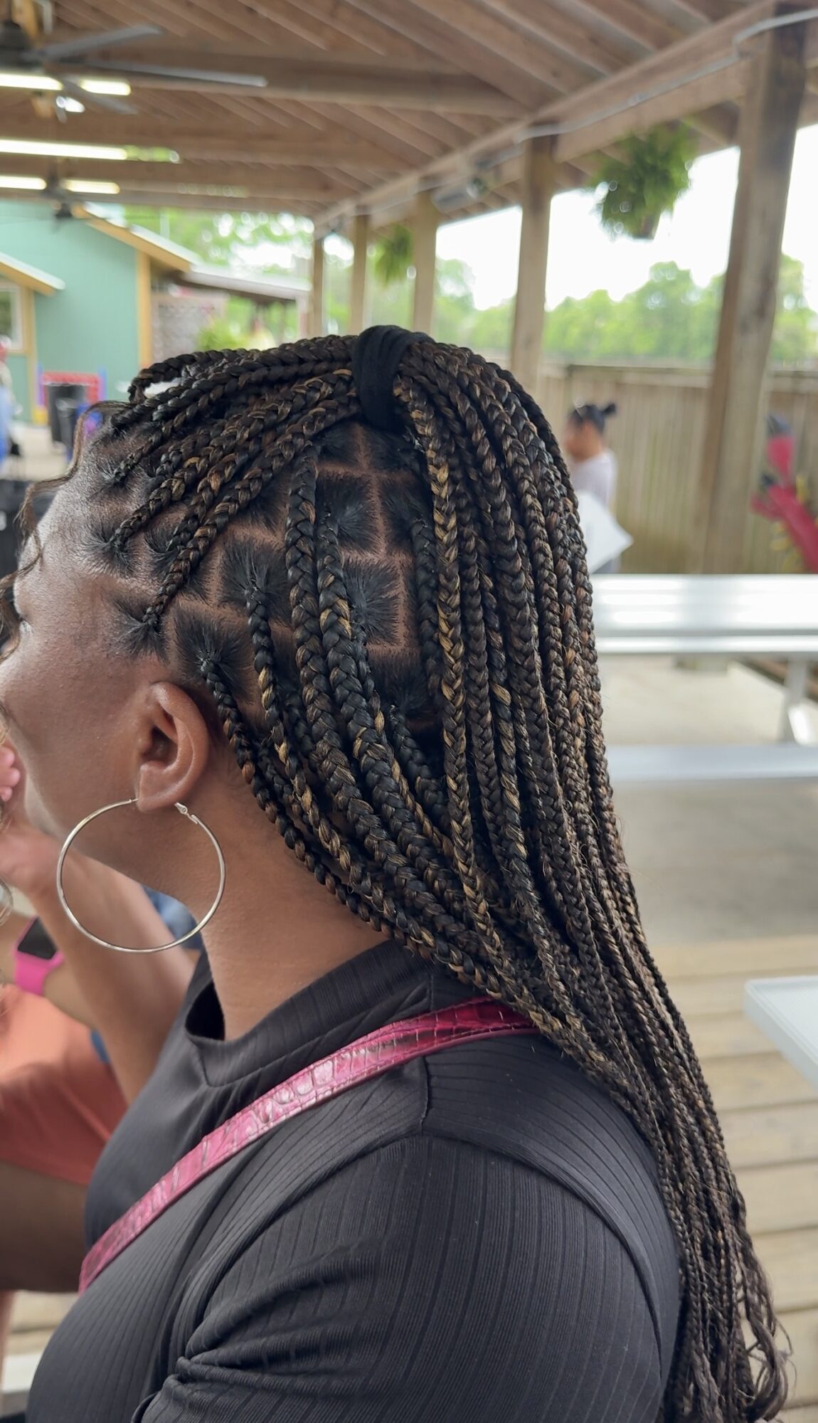 Side view of a woman with braided hair, wearing hoop earrings and a black top, outdoors under a wooden shelter.
