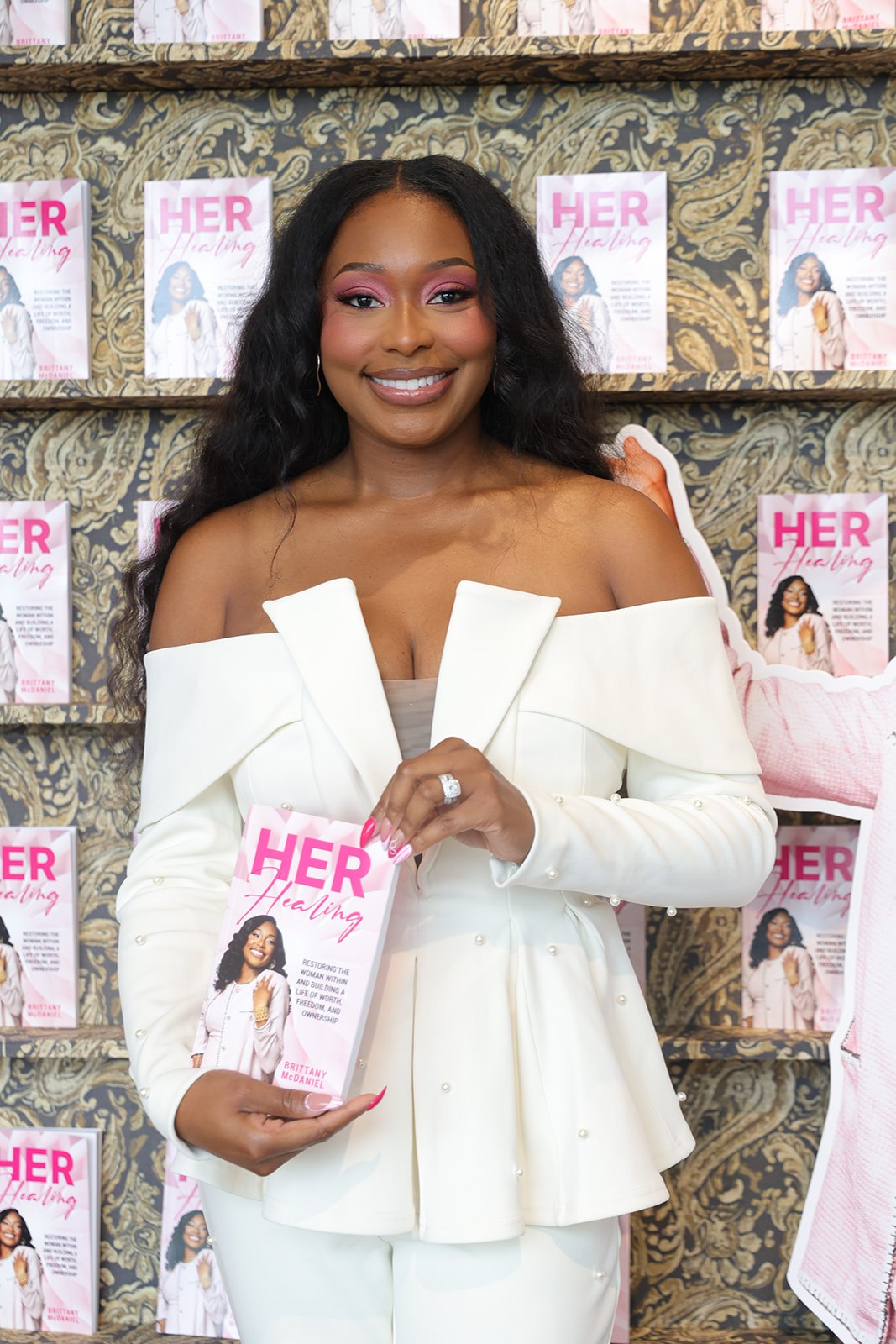 Smiling woman with long dark hair in white off-shoulder dress holding a magazine, standing in front of a wall with magazine covers.