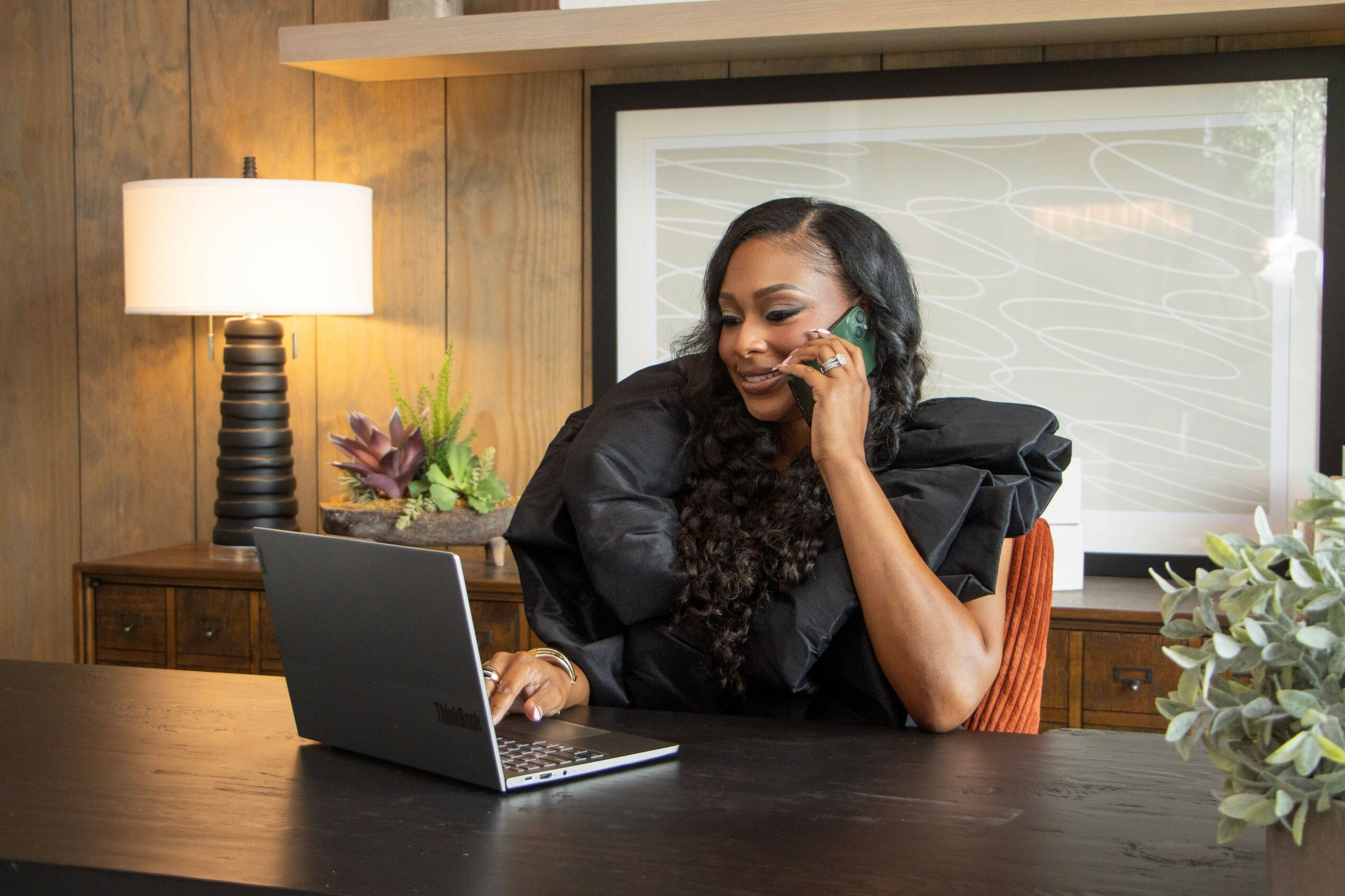 Woman with long curly hair sitting at a desk, smiling, with a laptop, in a room with wooden walls and a window.