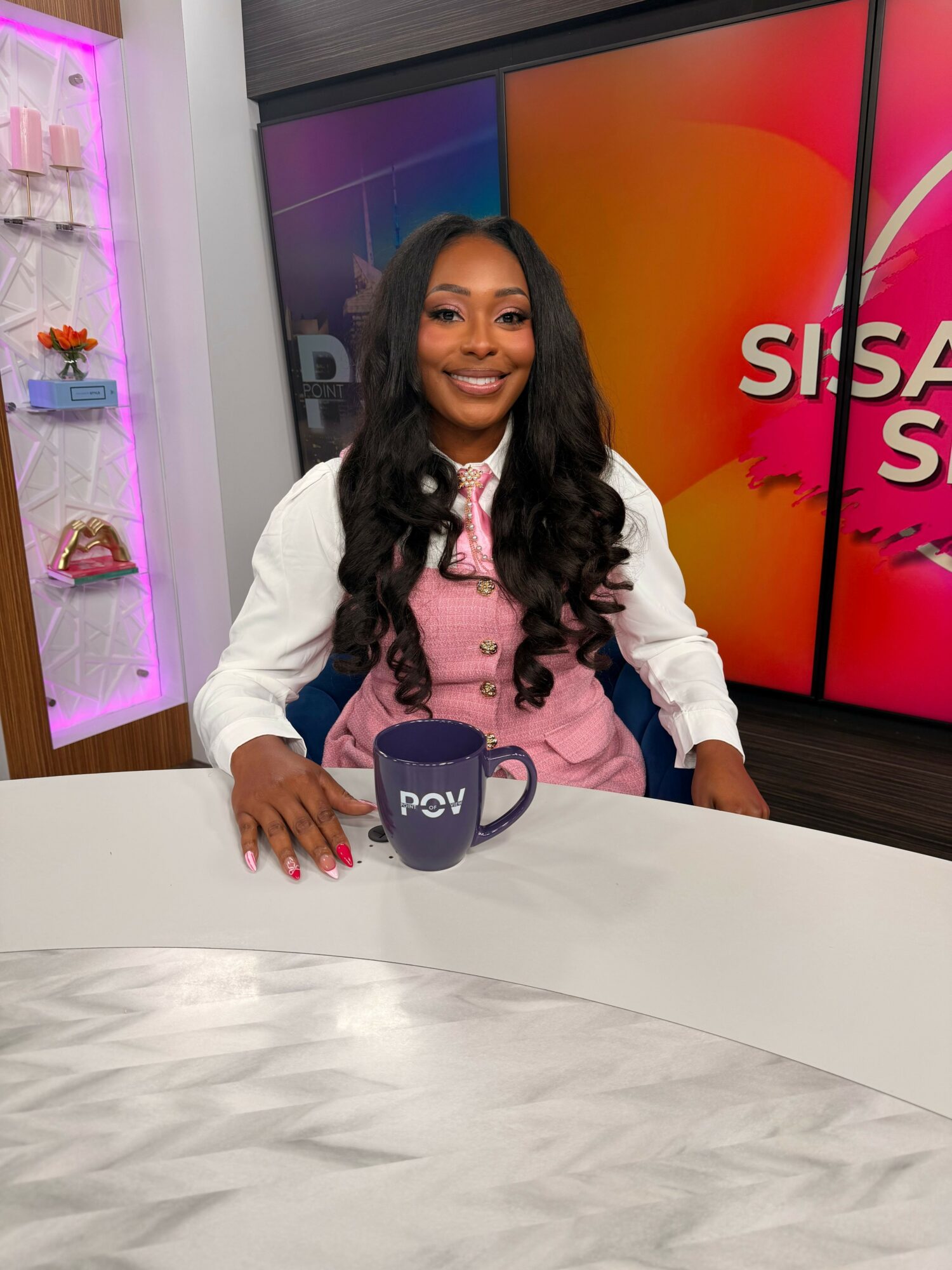 Young girl with long dark hair smiling at a table with a pink and purple background, holding a small bag.