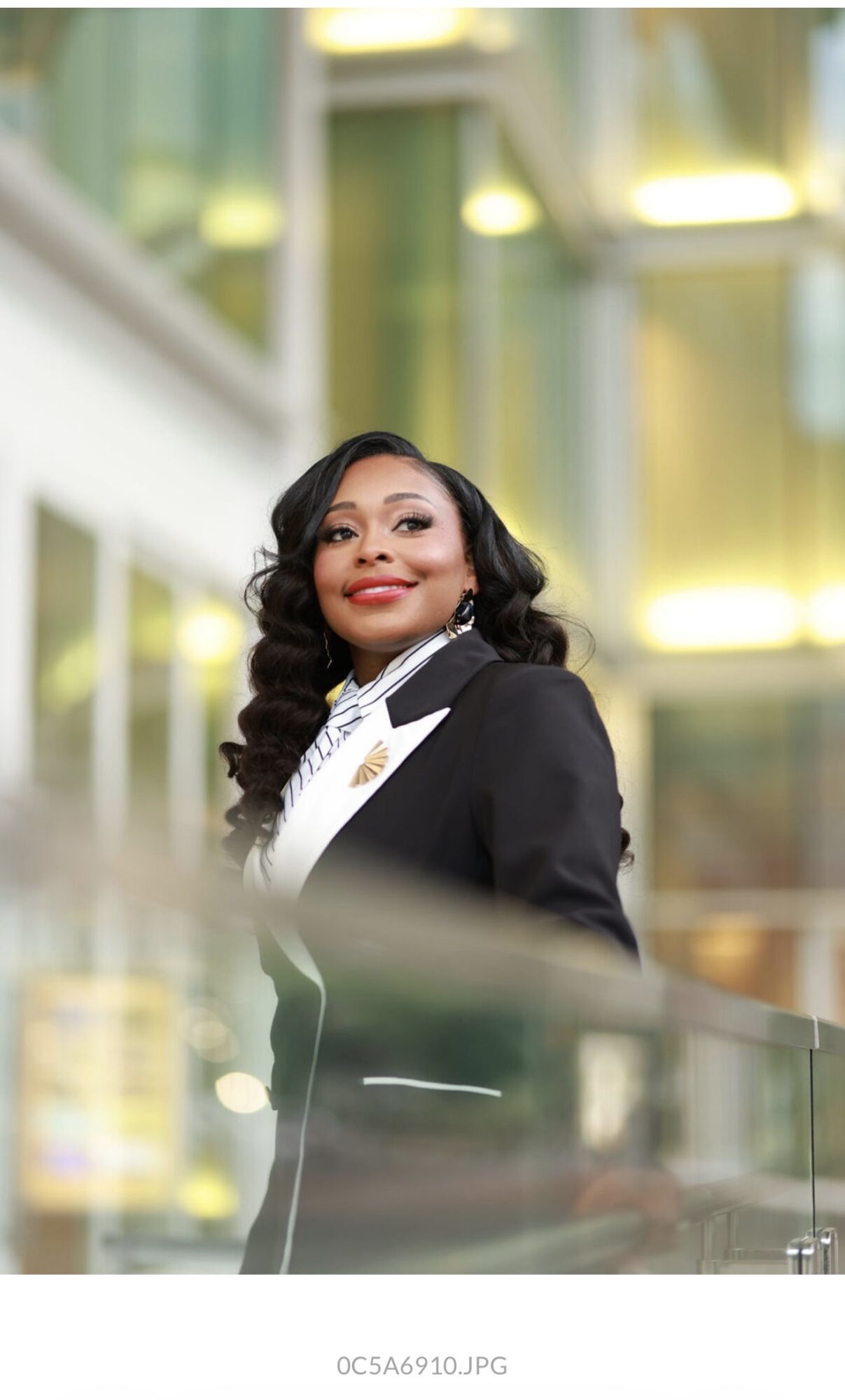 A woman with long dark hair and a black blazer stands indoors, smiling, with a modern building background.