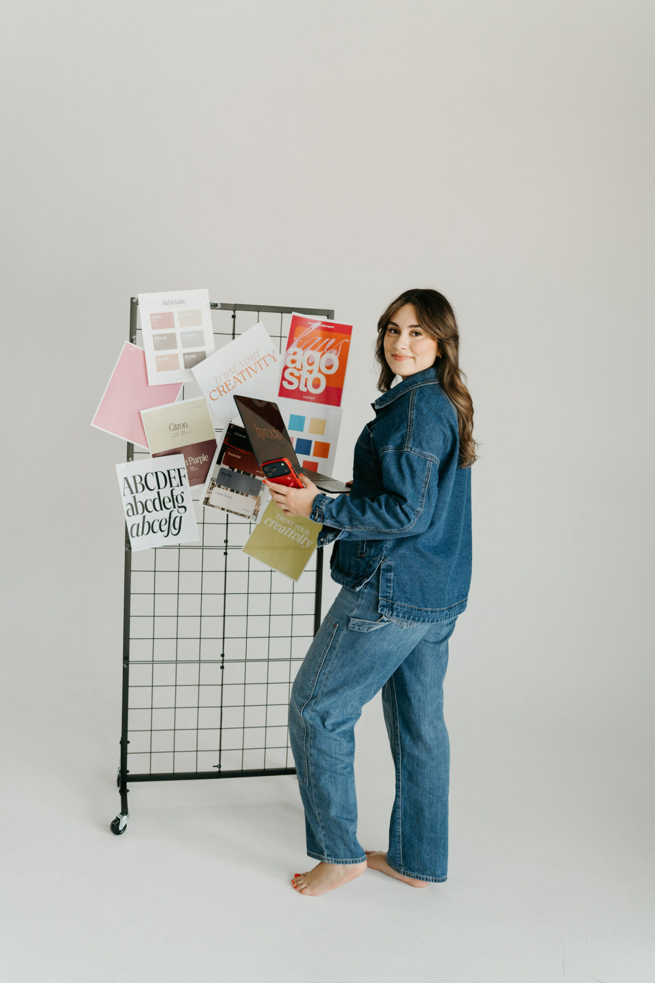 Woman with long hair in a denim jacket and jeans holds a laptop, standing next to a display with color samples and papers.
