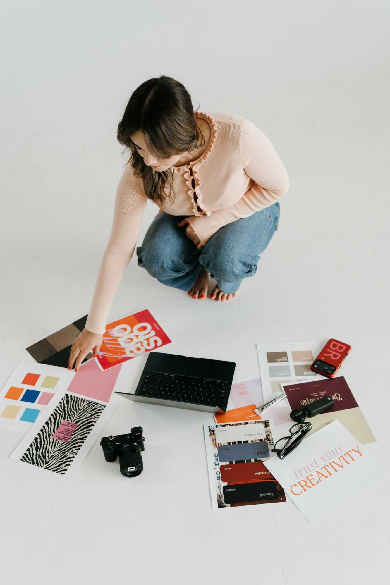 Person crouching, reaching for items on the floor, surrounded by color samples, a laptop, camera, and notebooks.