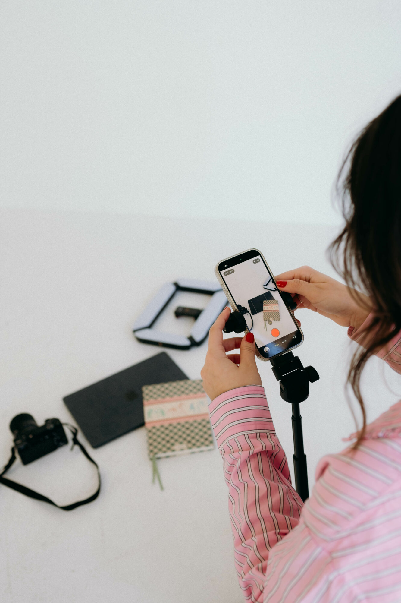 Person taking a photo with a smartphone mounted on a tripod, on a white table with camera equipment and a notebook