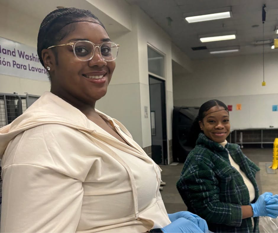 Two women wearing gloves in a room with white walls and fluorescent lights, smiling at the camera.