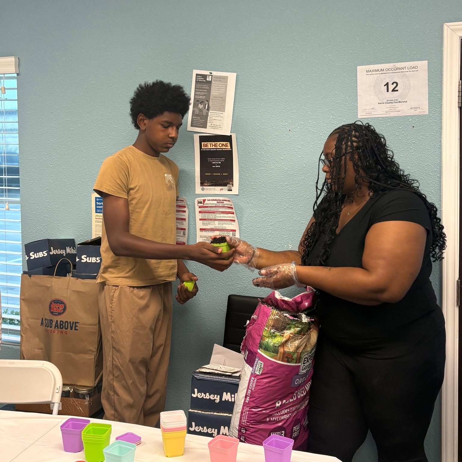 A young person and an adult woman exchange a small item in a room with boxes and posters on the wall.