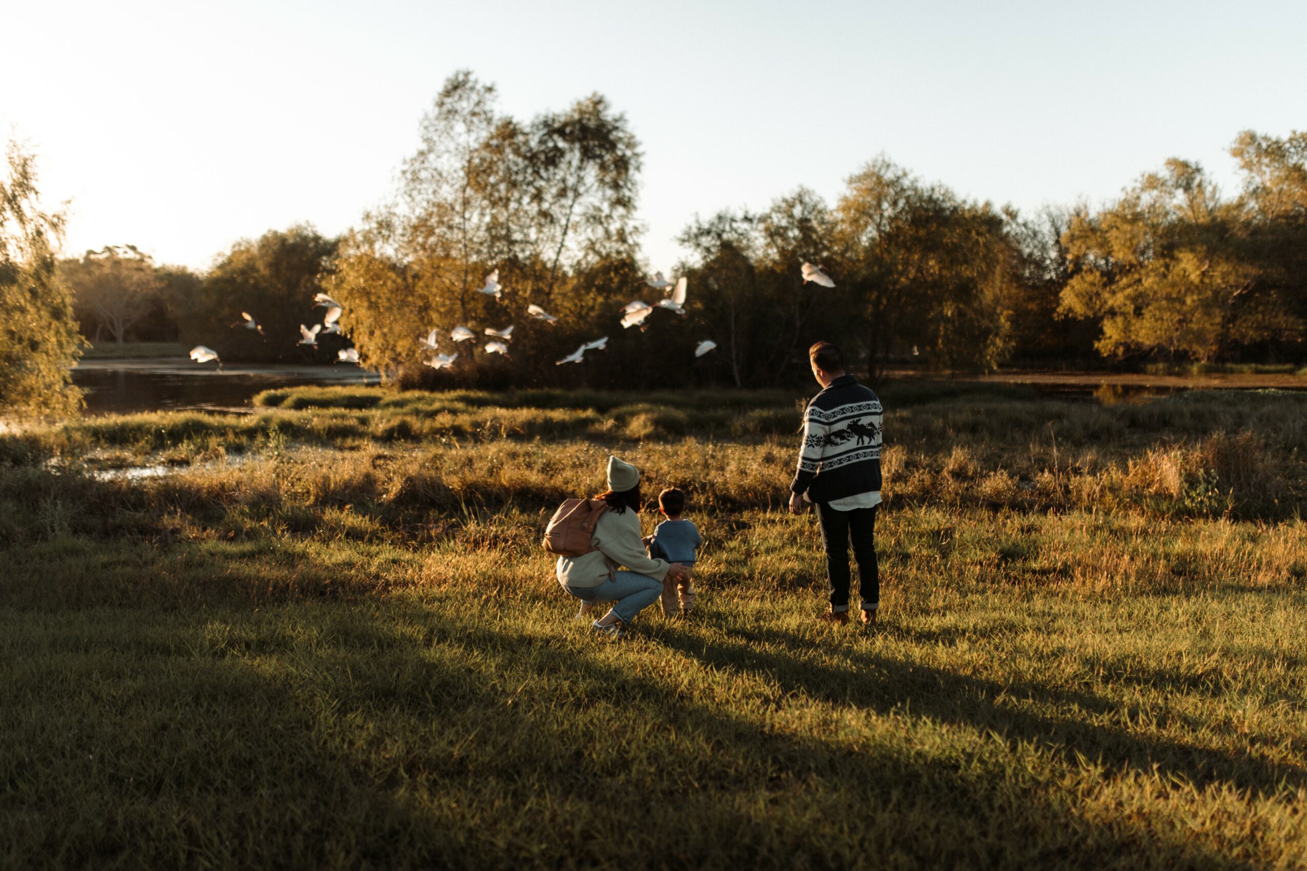 Person crouching with two children in a grassy field, another person standing nearby, trees in the background, sunlight.